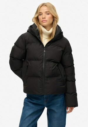 Blonde vrouw in een zwarte gewatteerde jas met capuchon, een crèmekleurige coltrui en een spijkerbroek, staande tegen een effen witte achtergrond.