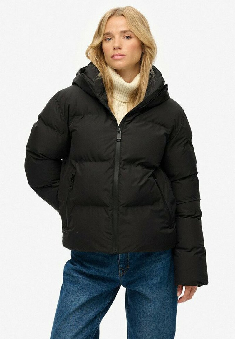 Femme blonde portant une doudoune noire avec capuche, un pull col roulé crème et un jean bleu, se tenant devant un fond blanc uni.