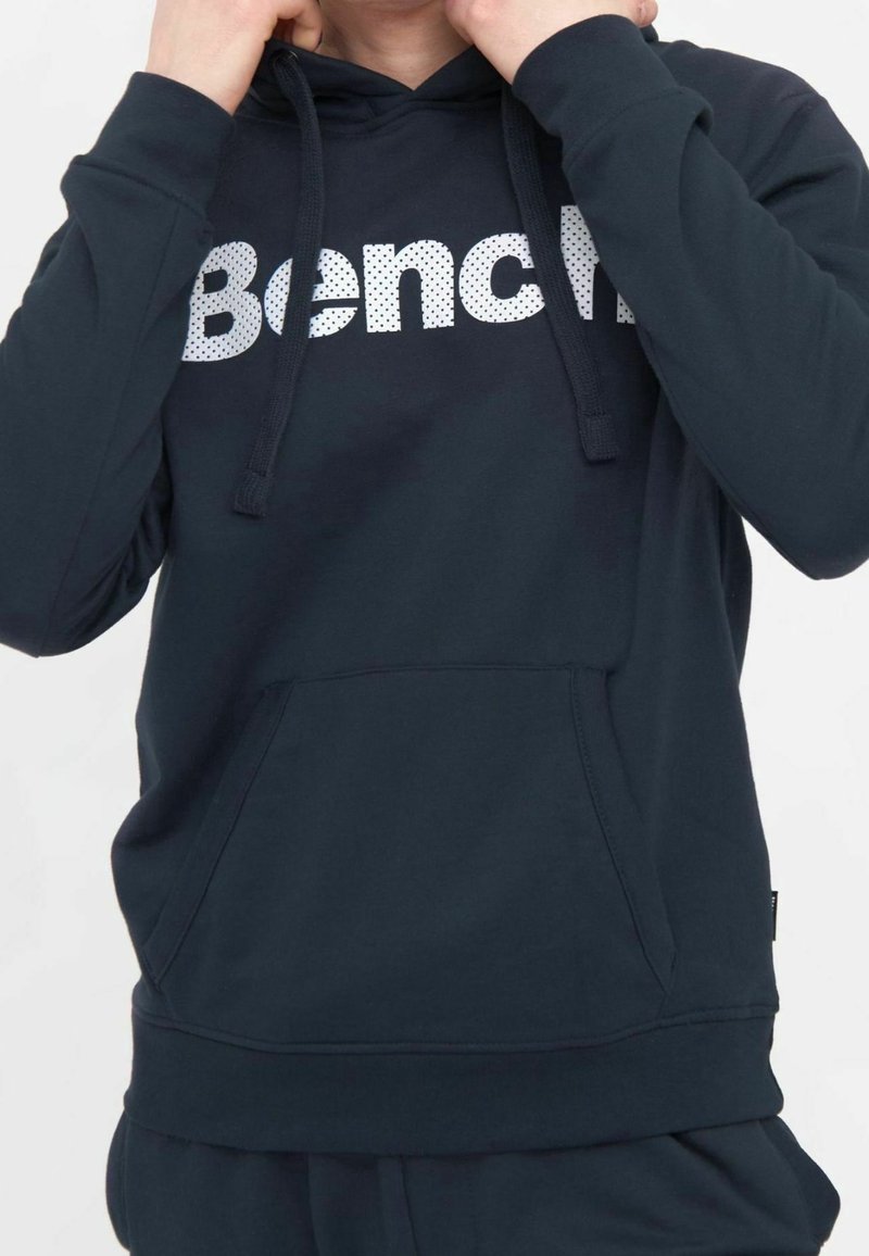 Sweatshirt marine en tissu doux avec une poche avant, capuche ajustable et un grand logo perforé blanc sur la poitrine.