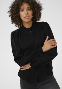 Musta paita, jossa on puolikorkea kaulus, napitus ja pitkät puff-hihat. Teksturoitu kangas, jossa on hillitty kiilto. Yhdistetty tummanharmaisiin farkkuihin.