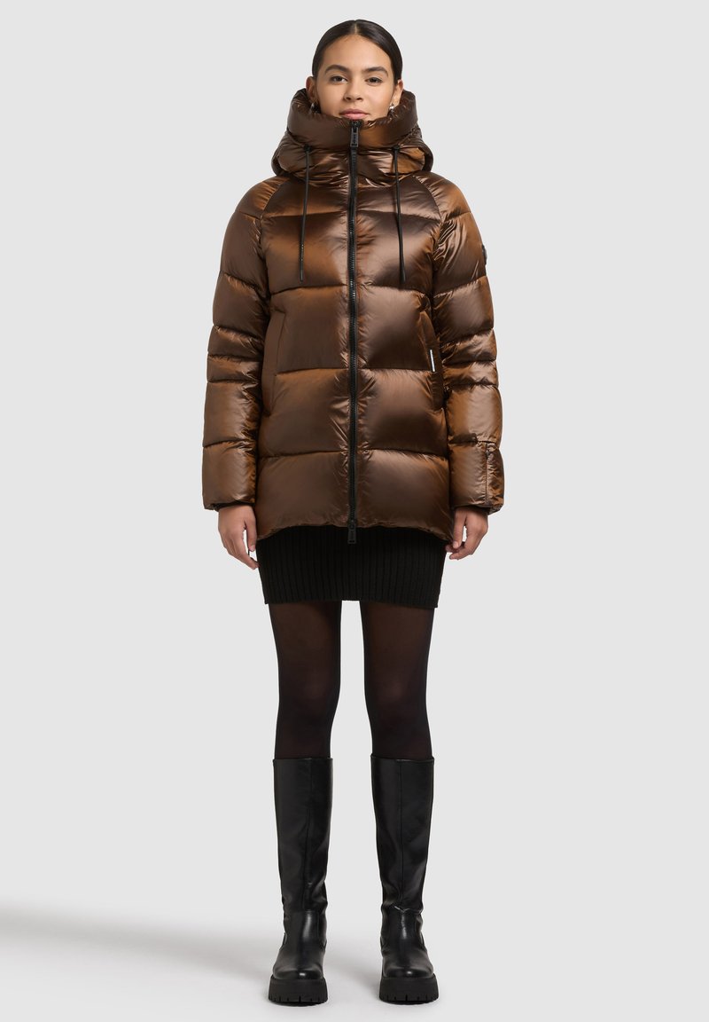 Brūna pūslāju jaka ar augstu apkakli un regulējamām auklām, ar rāvējslēdzēja aizdari un izšūtu rakstu, kopā ar melnu adītu svārku.