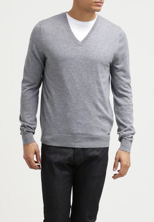 Mann trägt hellgrauen V-Ausschnitt-Pullover über weißem T-Shirt und schwarzen Hosen, steht vor schlichtem hellem Hintergrund.