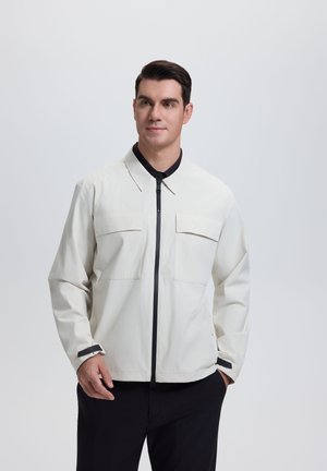 Mand iført en lysebeige jakke med sort lynlås og sorte manchetter, stående med den ene hånd i lommen mod en ensfarvet baggrund.