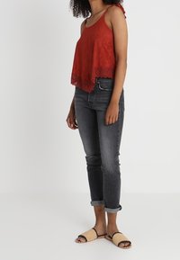 Rode mouwloze top met kanten rand, gecombineerd met donkere denim jeans. Model draagt beige gevlochten sandalen; comfortabele pasvorm, cropped stijl, zachte textuur.