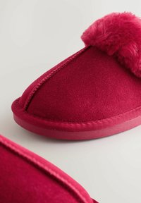 Piros papucs puha, bolyhos felsőrésszel, lekerekített formával és varrott részletekkel. Simább szövet külső rész és lapos gumitalp.