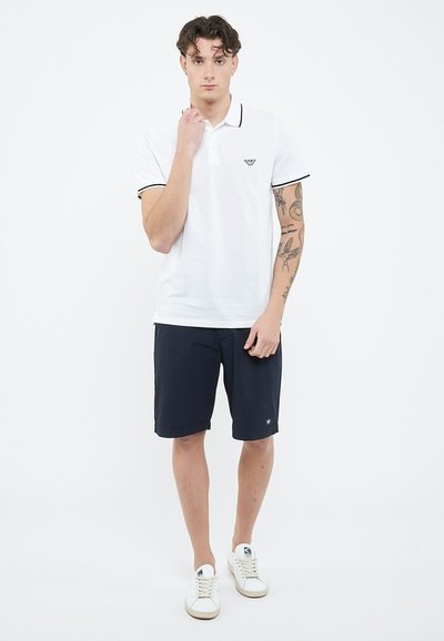 Młody mężczyzna w białej koszulce polo i czarnych krótkich spodenkach, stojący na tle białego tła, z tatuażami na prawej ramieniu.