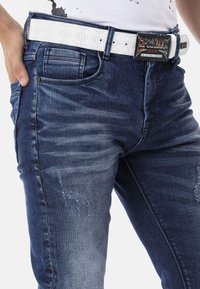 Jean en denim bleu foncé usé avec des motifs de décoloration claire, mettant en avant une ceinture texturée blanche et une boucle rectangulaire brillante.