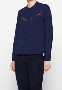 Camicia a maniche lunghe blu navy con chiusura a bottoni, colletto rotondo e dettagli in pizzo sulle spalle. Il tessuto ha un aspetto strutturato e spiegazzato.