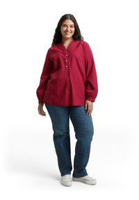 Rotes Langarmshirt mit V-Ausschnitt und Knopfdetails, kombiniert mit blauen Jeans und weißen Sneakern. Das Shirt zeichnet sich durch einen strukturierten Stoff aus.