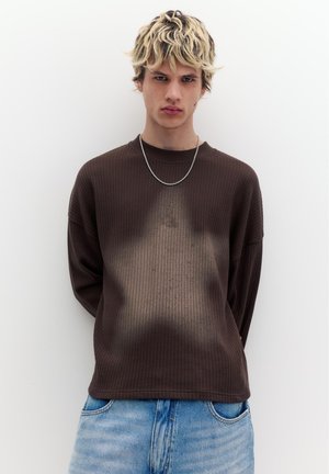 Junger Mann mit blondem Haar, der einen dunkelbraunen strukturierten Pullover mit Sternmuster und hellblaue Jeans trägt, steht vor einem weißen Hintergrund.