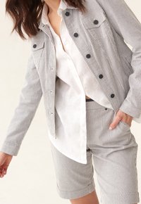 Marynarka z paskowanym materiałem i czarnymi guzikami, połączona z białą koszulą oraz dopasowanymi krótkimi spodniami w paski. Posiada luźny krój i kieszenie z przodu.