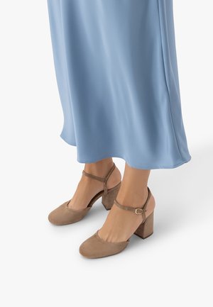 Stopy w beżowych zamszowych butach na słupku z paskami wokół kostki, stojące na białej powierzchni, w jasnoniebieskiej, zwiewnej spódnicy.