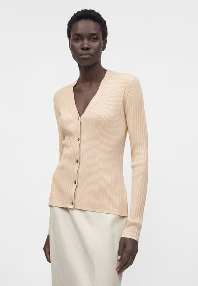 Γυναίκα με σκούρο δέρμα, που φοράει μπεζ ριπ ζακέτα με κουμπιά και ανοιχτό κρεμ χρώμα φούστα, στέκεται μπροστά σε ένα απλό λευκό φόντο.