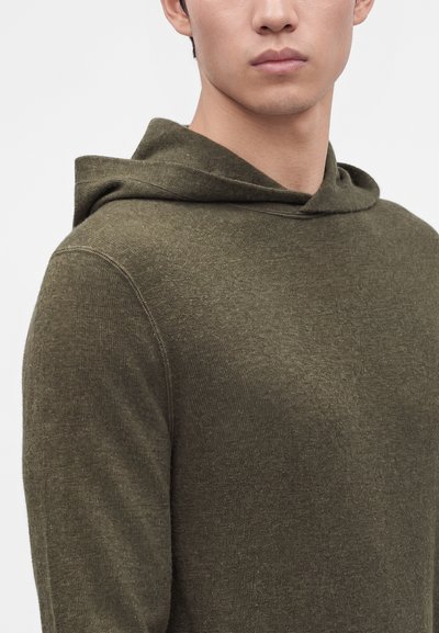 Νέος άνδρας που φοράει πλεκτό hoodie σε ελαιί πράσινο χρώμα, εμφανιζόμενος από τους ώμους και πάνω με φόντο ένα απλό ανοιχτό χρώμα.