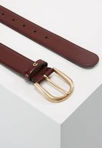 Leren riem in donkerburgundy met een gladde afwerking. Voorzien van een grote gouden gesp en meerdere gaatjes voor maatverstelling.