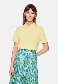 Gelbes kurzärmeliges Poloshirt mit Kragen und vier Knöpfen, kombiniert mit einem blau-grünen gemusterten Rock mit fließenden Pinselstrich-Designs.