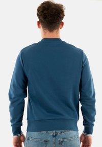 Felpa blu a girocollo in materiale morbido, con polsini e orlo ribattuti e sottili accenti a righe sui polsini. Vista posteriore semplice.