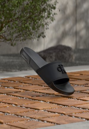 Mule noire Calvin Klein avec logo CK en relief, exposée en extérieur sur des dalles en bois avec du gravier entre les interstices, feuillage vert flou en arrière-plan.