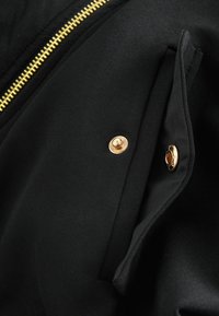 Sort stofjakke med en fremtrædende guld lynlås, trykknapsdetaljer og et elegant lomme-design. Glat tekstur, struktureret form.