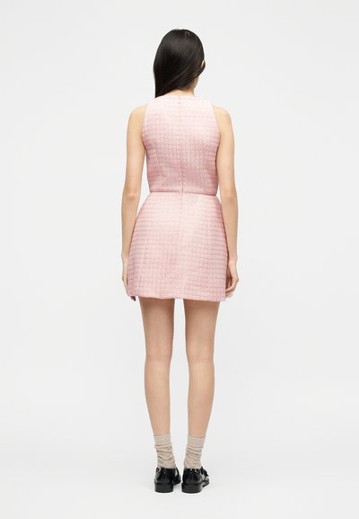 Γυναίκα με μακριά σκούρα μαλλιά που φορεί ροζ, μίνι φόρεμα χωρίς μανίκια με υφή, μπεζ κάλτσες και μαύρα μοκασίνια, όρθια με την πλάτη προς τον θεατή.