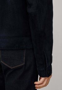 Marineblauwe sued jacket met een gladde textuur, een getailleerde pasvorm en knoopdetails bij de manchetten. Gecombineerd met donkere jeans van denim met contrasterende stiksels.
