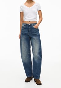 Mujer con una camiseta blanca de cuello en V corta, jeans azules de cintura alta y holgados, y botas marrones hasta el tobillo, de pie con una mano en el bolsillo.