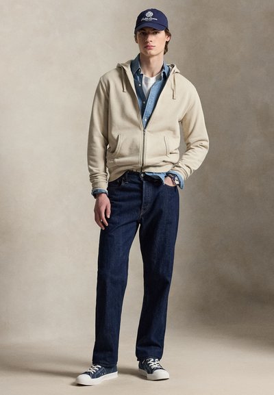 Young man dressed in beige zip-up hoodie over blue denim shirt and white tee, dark jeans, navy sneakers, and blue baseball cap.