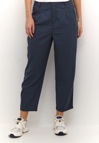 Pantalons bleu marine avec une coupe ajustée, des plis avant et un design court, associés à des chaussures de sport blanches et grises.