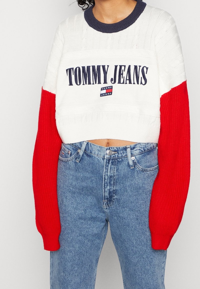 Ściągaczowa bluzka w kolorze białym, czerwonym i granatowym o dzianinowej fakturze. Z przodu znajduje się logo "TOMMY JEANS". W komplecie z dżinsami z wysokim stanem w kolorze niebieskim.