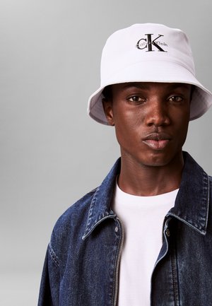 Giovane uomo che indossa un cappello bucket bianco Calvin Klein, una camicia bianca e una giacca di jeans blu su uno sfondo grigio uniforme.