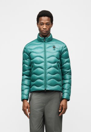 Mladić u tirkiznoj prošívanoj jakni sa patentnim zatvaračem i sivim hlačama, stoji nasuprot jednobojnoj bijeloj pozadini.