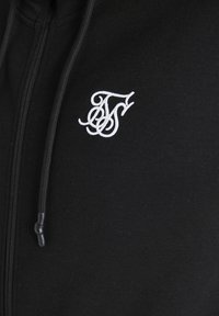 Sudadera con cremallera de color negro hecha de un tejido suave. Presenta un logo bordado en blanco en el pecho y cordones negros en la capucha.
