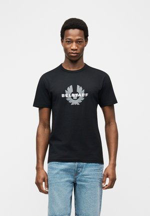 Mladý muž nosící černé tričko s logem Belstaff a světlé modré džíny, stojící před jednobarevným bílým pozadím.