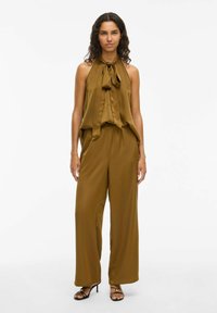Jumpsuit in satin marrone con scollo a halter e nodo, gambe larghe e vestibilità rilassata. Abbinato a sandali neri con tacco alto e dettagli dorati.