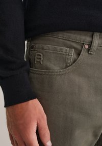 Jeans verde olivo con un acabado texturizado, que presentan un bolsillo trasero con una letra "A" en relieve y detalles de botones metálicos. Suéter de punto negro visible.