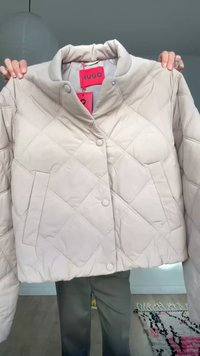 Világos bézs színű, steppelt dzseki patentgombokkal és oldalzsebekkel, beltéren szürke nadrágot viselő személy által tartva.