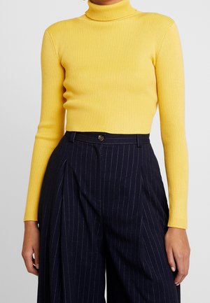 Femme portant un pull à col roulé jaune côtelé rentrée dans un pantalon large noir à taille haute à fines rayures.