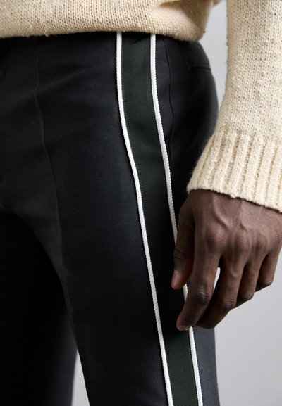 Zwarte broek met een groene zijstreep en witte bies. De stof lijkt glad. Een gebreide beige trui is gedeeltelijk zichtbaar.