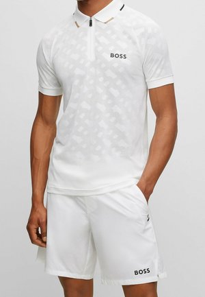 Mann trägt weißes BOSS-Poloshirt mit dezentem Muster und weiße BOSS-Shorts, eine Hand in der Tasche, vor einfarbigem Hintergrund.