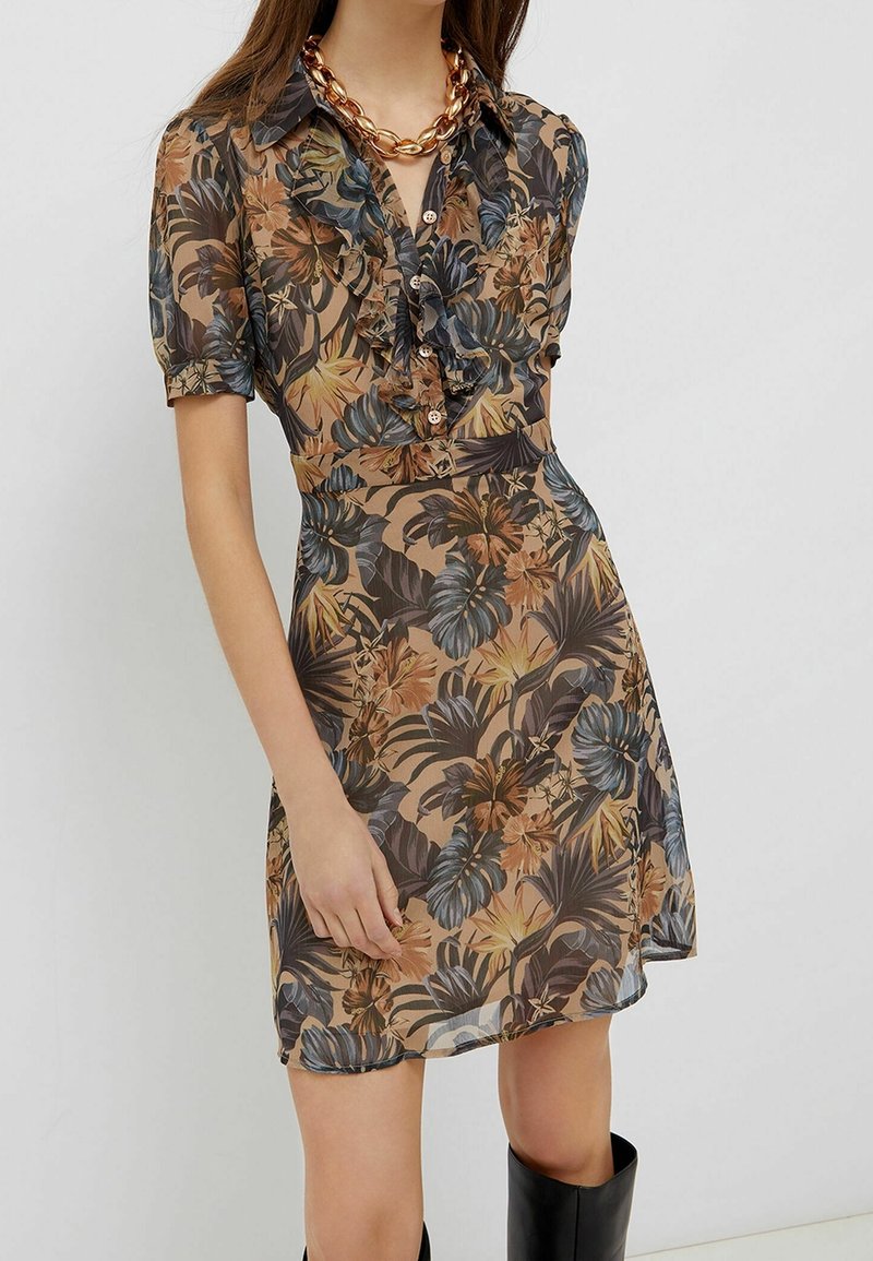 Femme portant une robe fleurie à manches courtes, boutonnée, à volants, dans des tons marron, noir et orange, accessoirisée d'un collier chaîne en or massif.
