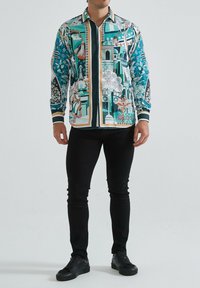 Chemise à boutons avec des motifs complexes teal et blancs, présentant des designs floraux et architecturaux. Manches et col noirs contrastants.