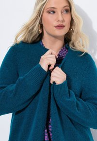 Teal stickad cardigan med ribbade ärmslut och öppen front, lagd över en lila prickig topp. Mjuk textur och medelvikt.