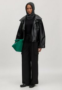 Schwarze Leder-Übergrößejacke, eng anliegendes schwarzes Top, weite schwarze Hose, grüne Tasche, grauer Strickkopftuch und schwarze Schuhe mit Fransen-Detail.