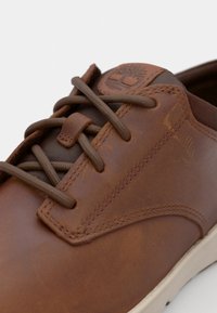 Zapato de cuero marrón con una textura suave, cordones oscuros contrastantes y un logo en la lengüeta. Incluye una suela de goma beige.