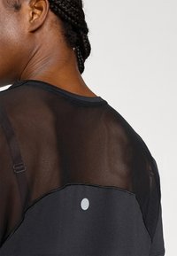 Μαύρο αθλητικό τοπ με ημιδιάφανο πλέγμα στην πλάτη, στρογγυλή λαιμόκοψη και μικρή αντανακλαστική λεπτομέρεια κοντά στη ραφή του ώμου.