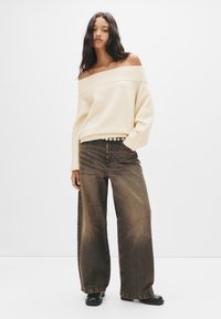 Pullover crema off-the-shoulder con collo a costine, abbinato a jeans denim marrone chiaro a gamba larga e stivaletti neri alla caviglia.