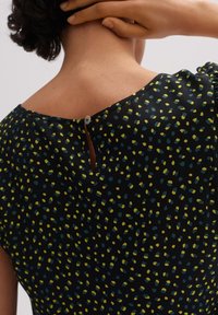 Top a maniche corte di colore nero con motivo, caratterizzato da macchie sparse gialle e turchesi, con apertura a occhiello e dettaglio di bottone sul retro del collo.