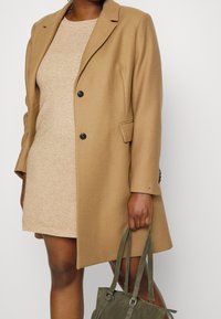 Manteau beige en mélange de laine avec revers crantés, deux boutons et poches latérales, associé à une robe texturée beige clair et un sac à main vert olive.