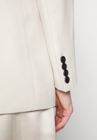 Manche de blazer beige avec quatre boutons noirs près du poignet, portée avec un pantalon beige assorti, main visible sur le côté.