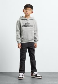 Pelēka kapa jaka ar "Nike Sportswear" grafiku, priekšējā kabatā, kombinēta ar melniem biksēm un melnbaltām/sarkanām kedām, stāv uz pelēka virsmas.